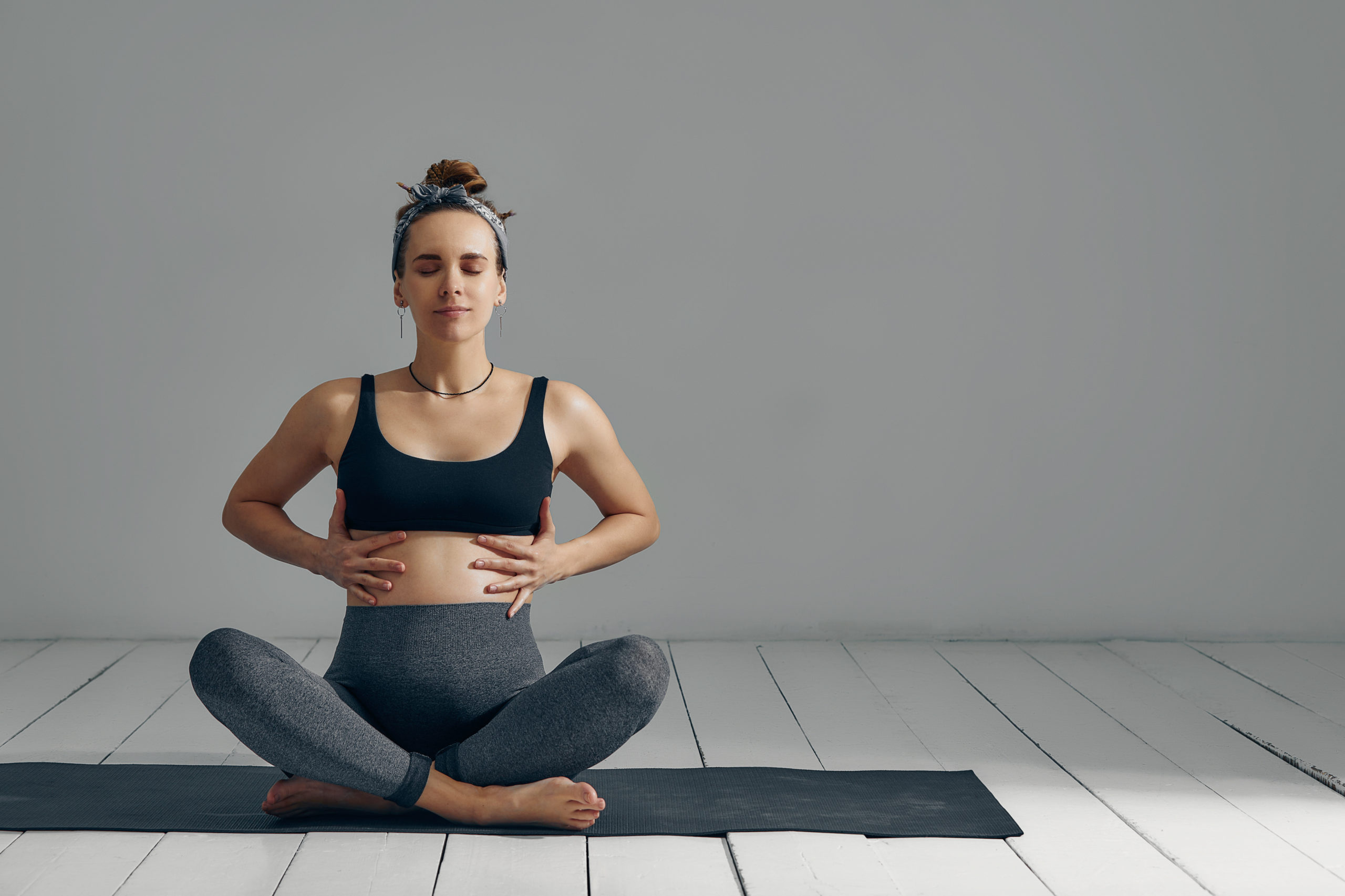 Young pregnant millennial woman sitting on the mat touches her belly after performing prenatal and meditation exercises at a yoga class - Concept of life and maternity